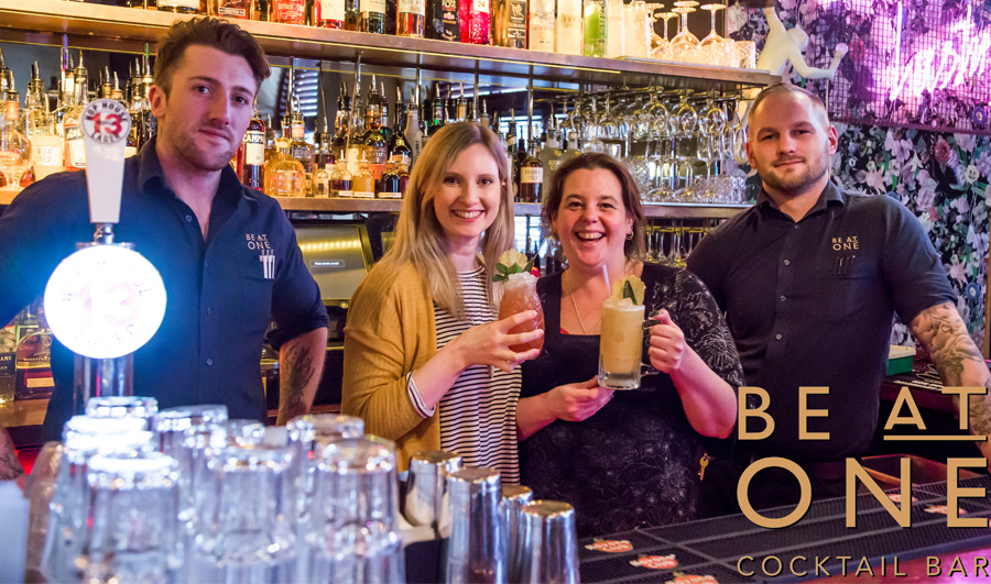 Four people standing behind a bar holding cocktails
