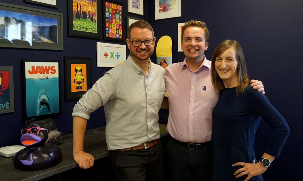 Aimee, Alex and James standing in front of picture wall